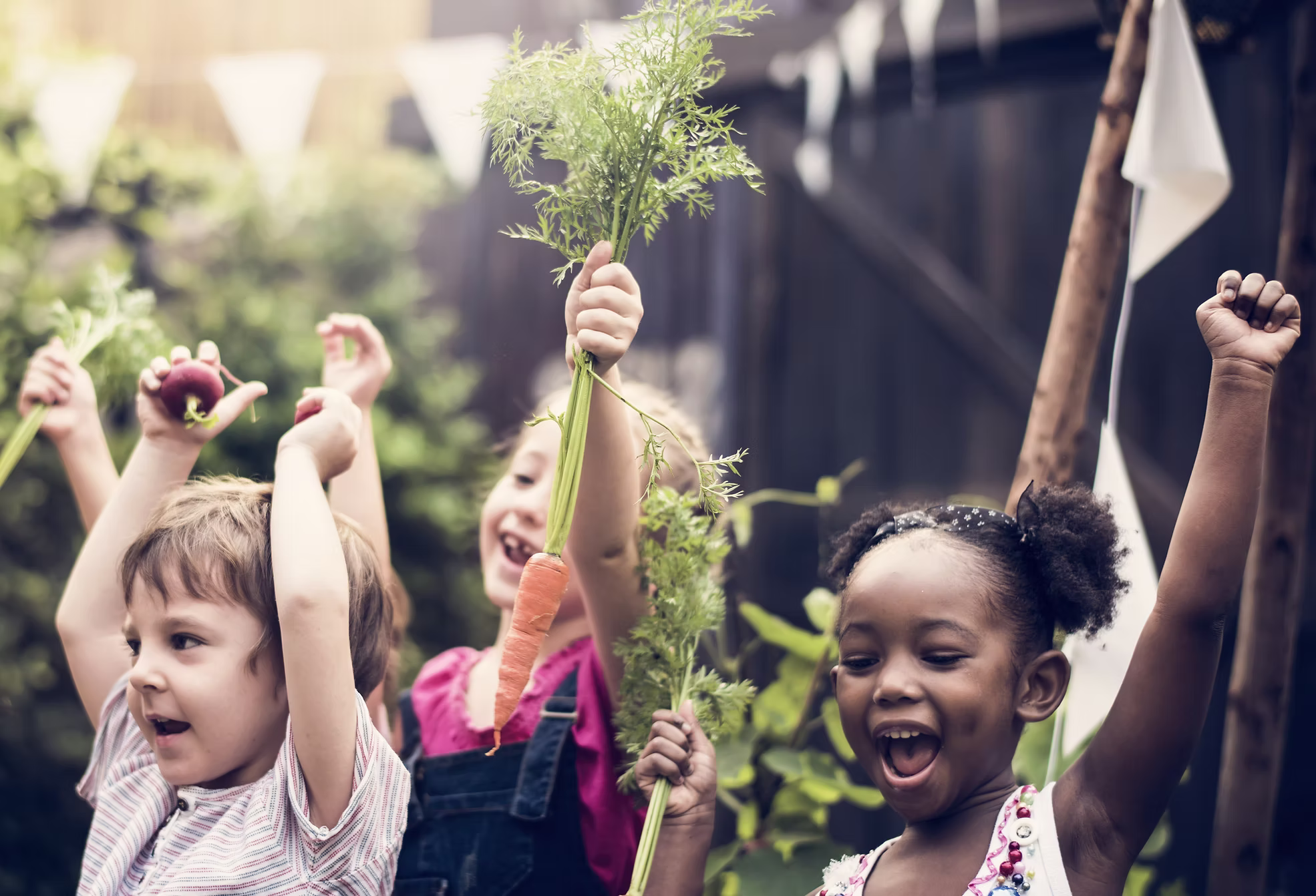 Drei glückliche Kinder halten in einem Garten Karotten am Suppengrün hoch